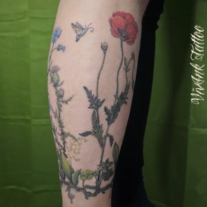 Színes, realisztikus, virágos tetoválás lábszáron - pipacs, gyöngyvirág, bogáncsvirág, kecskefarkú szender